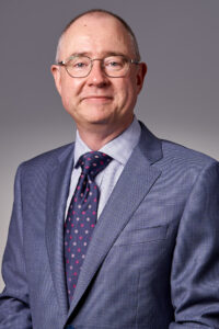 Mark de Raad is a man in his fifties with short dark hair, round glasses and is wearing a grey suit.