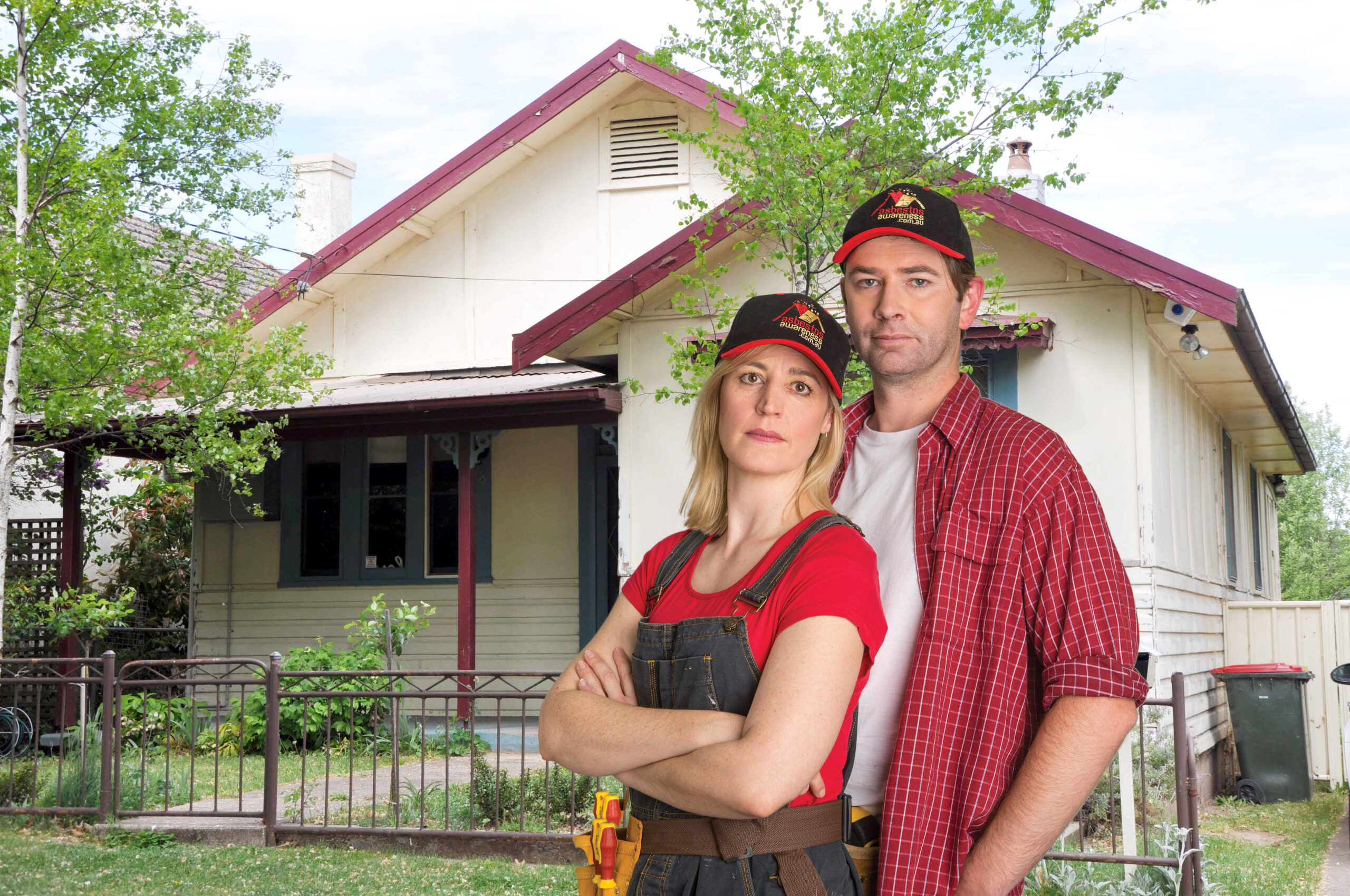Couple wearing renovating clothes and Asbestos Awareness hats stand in front of hold home with stern expressions.
